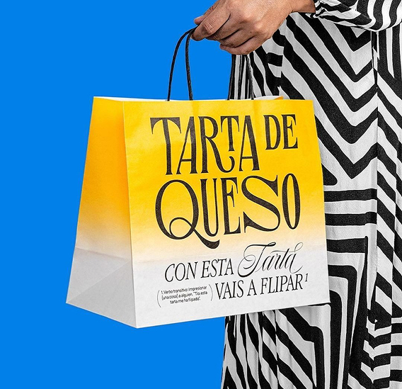 Persona sosteniendo una bolsa de papel amarilla y blanca con asas negras y el texto “TARTA DE QUESO”, sobre fondo azul.