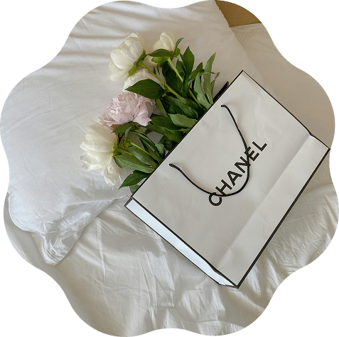 Bolsa de papel blanca de CHANEL con asas negras junto a un ramo de flores sobre una cama con sábanas blancas.