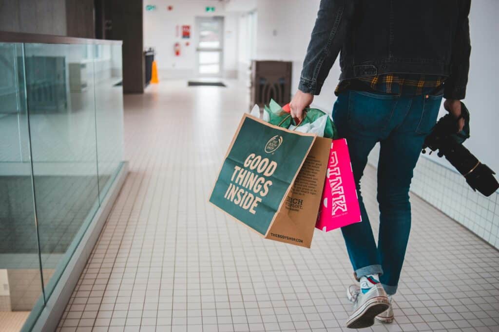 Persona caminando por un centro comercial con varias bolsas de compras de marca y una cámara en la mano; destaca una bolsa verde con el texto “GOOD THINGS INSIDE”.