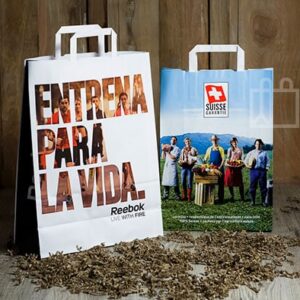 Bolsas de papel personalizadas con asa plana: una de Reebok con el texto “ENTRENA PARA LA VIDA” y otra de Suisse Cheese con imagen de granjeros, sobre fondo de madera.