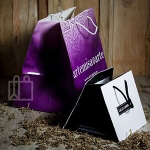 Bolsas de papel personalizadas: una morada con asas blancas y texto “artemisa&arte” y otra negra con bolsa blanca con logo “M”, sobre fondo de madera.