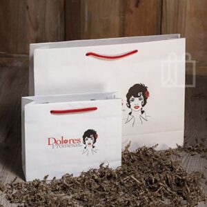 Bolsas de papel blancas personalizadas con asas rojas y logo “Dolores Promesas”, sobre fondo de madera con relleno decorativo.